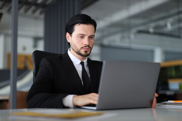A man in a suit is sitting at a desk with a laptop in front of him. He is focused on his work, possibly typing or browsing the internet. Concept of professionalism and productivity
