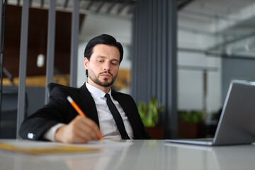 A man in a suit is writing with a pencil on a desk. Concept of professionalism and focus, as the man is likely working on a task or project. The presence of the laptop