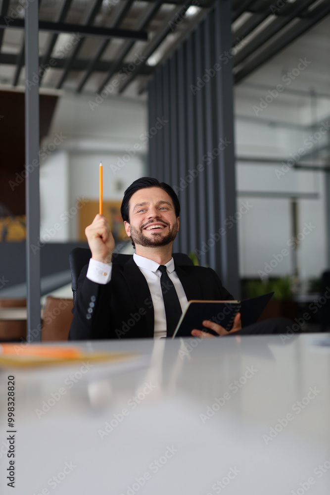 Poster a man in a suit is holding a pencil and smiling. he is sitting at a desk with a notebook and a book.