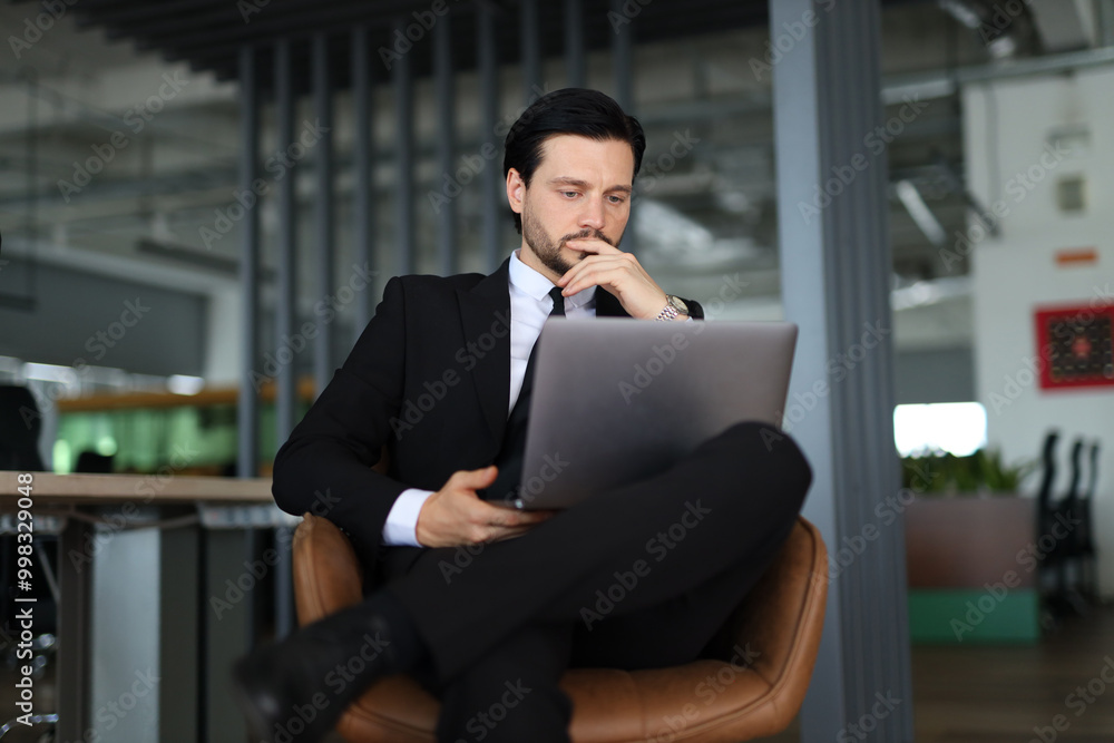 Poster a man in a suit is sitting in a chair with a laptop in front of him. he is deep in thought, possibly