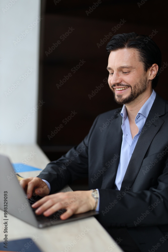 Poster a man in a suit is sitting at a table with a laptop in front of him. he is smiling and he is enjoyin