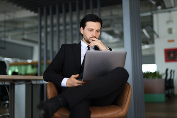 A man in a suit is sitting in a chair with a laptop in front of him. He is deep in thought,...