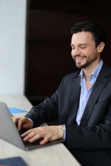 A man in a suit is sitting at a table with a laptop in front of him. He is smiling and he is enjoying himself