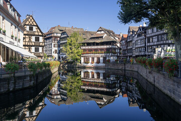 Naklejka premium Strasbourg, France - August 28th 2024 : View of the Tanner's Quarter with half-timbered buildings and the Ill river and reflections on the water.