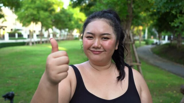 Tired overweight asian woman sweating after exercise and resting and show thumbs up in Garden outdoors . fat female break workout outside in morning