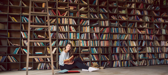 girl sitting on the floor in big library with many bookshelves at the background. Happy smiling weekend hobby time. Long horizontal banner atmospheric interior