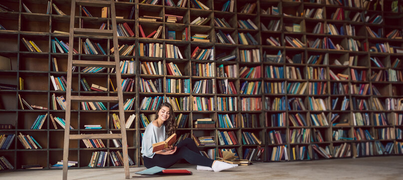 girl sitting on the floor in big library with many bookshelves at the background. Happy smiling weekend hobby time. Long horizontal banner atmospheric interior
