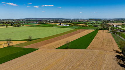 Expansive fields in different shades of green and brown create an eye-catching patchwork in a serene rural area. Bright blue skies enhance the tranquil atmosphere, highlighting agricultural activity.