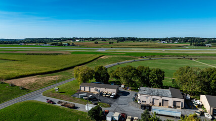 The aerial perspective captures a rural airport with a visible runway, surrounded by fields and greenery. Nearby buildings and vehicles create a sense of activity under a clear blue sky.