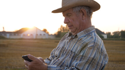 Portrait elderly old caucasian man farmer agronomist worker in agricultural field at sunset. Director using phone smartphone tablet, checking harvest crop. Against village and farm with warehouse