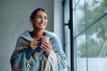 Happy woman wrapped in blanket drinking tea by  window.