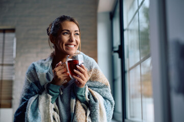 Happy woman wrapped in  blanket enjoying in cup of warm tea at home.