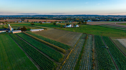 The sun sets over a picturesque rural landscape, highlighting expansive fields of crops and farmhouses scattered throughout. The vibrant green and brown layers form a patchwork effect. © Greg Kelton