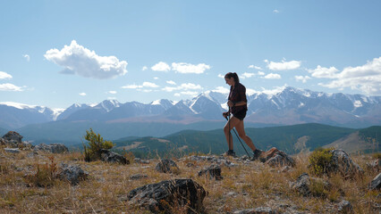 Active free athletic woman tourist blogger hiking in valley with trekking poles outdoors on sunny day on rocks of hills. Background of snow capped summit of mountains. Summer travel holiday nature