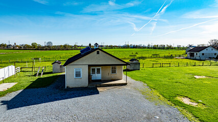 Fototapeta premium An Amish one room school house sits on a gravel area, surrounded by expansive green fields, barns, and distant structures under a bright blue sky.