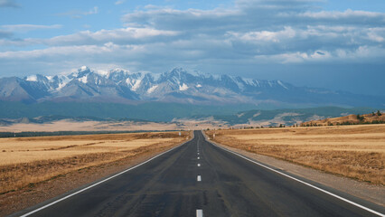 Driving on highway of black asphalt and white road markings at sunny day, road going into distance through desert prairie at autumn to snowy mountain summits on horizon in wild. Pov from moving car