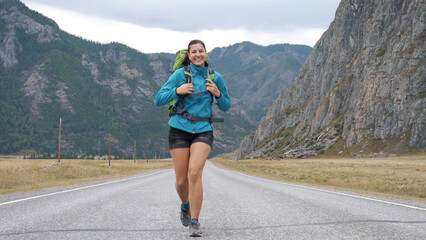 Happy free caucasian woman with backpack running to meet camera smiling. Positive emotions from meeting after separation. Running on asphalt road against the of rocky mountains. Slow motion