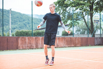 Young man casually juggles a football on an outdoor sports court, focusing on his footwork. Dressed in casual sportswear, he practices ball control in a vibrant natural setting