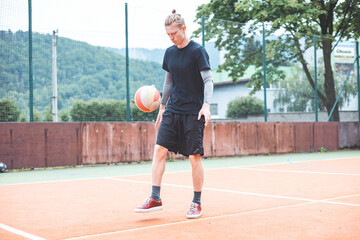 Young man casually juggles a football on an outdoor sports court, focusing on his footwork. Dressed in casual sportswear, he practices ball control in a vibrant natural setting