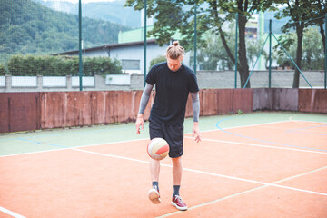 Young man casually juggles a football on an outdoor sports court, focusing on his footwork. Dressed in casual sportswear, he practices ball control in a vibrant natural setting