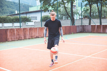 Young man casually juggles a football on an outdoor sports court, focusing on his footwork. Dressed in casual sportswear, he practices ball control in a vibrant natural setting