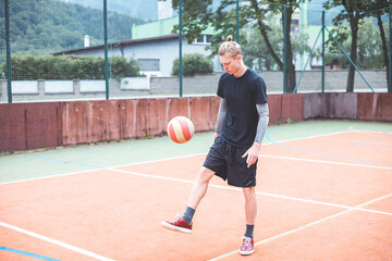Young man casually juggles a football on an outdoor sports court, focusing on his footwork. Dressed in casual sportswear, he practices ball control in a vibrant natural setting