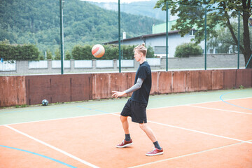 Fototapeta premium Young man with a man bun juggles a football on a vibrant orange court during a sunny day. Wearing casual sportswear, he focuses on controlling the ball with precision in a natural setting