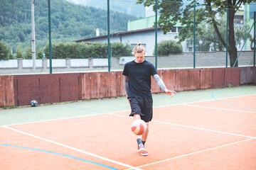 Young man with a man bun juggles a football on a vibrant orange court during a sunny day. Wearing casual sportswear, he focuses on controlling the ball with precision in a natural setting