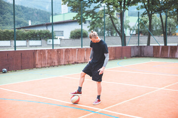 Young man with a man bun juggles a football on a vibrant orange court during a sunny day. Wearing casual sportswear, he focuses on controlling the ball with precision in a natural setting