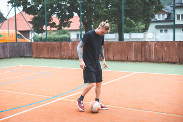 Fototapeta premium Young man with a man bun juggles a football on a vibrant orange court during a sunny day. Wearing casual sportswear, he focuses on controlling the ball with precision in a natural setting
