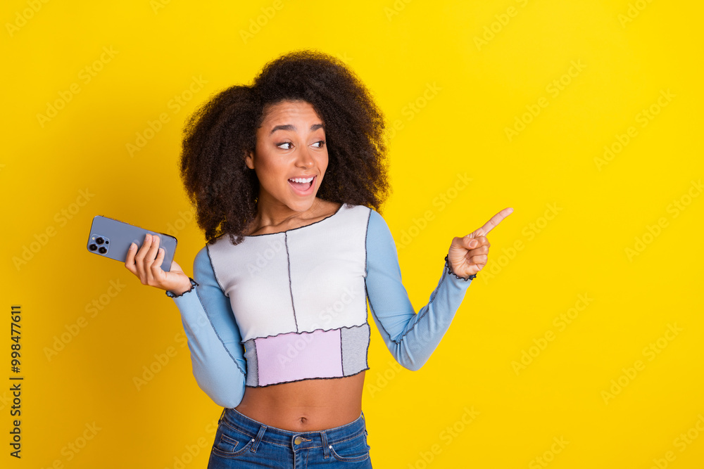 Wall mural Photo of attractive young woman hold gadget point look empty space dressed stylish blue clothes isolated on yellow color background