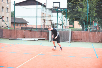 Young man focuses on controlling a football on an outdoor court. He carefully juggles the ball with precision, surrounded by an urban backdrop of buildings and a goal, combining sports and skill