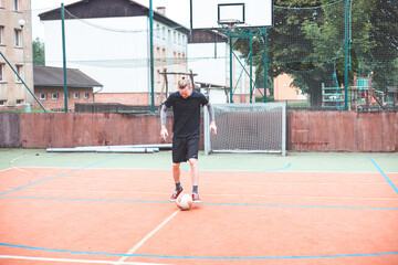 Young man focuses on controlling a football on an outdoor court. He carefully juggles the ball with precision, surrounded by an urban backdrop of buildings and a goal, combining sports and skill