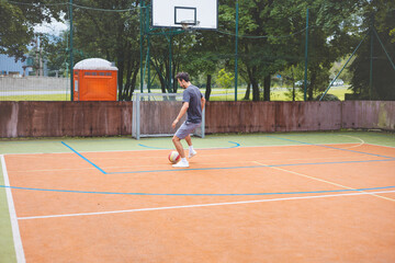 Young man dribbles a football with precision on an outdoor court, practicing his footwork in a casual environment. The court is surrounded by nature, providing a peaceful backdrop