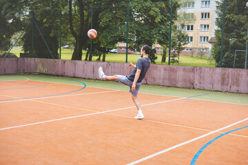 Young man takes a powerful shot on goal while playing football on an outdoor court. Wearing casual attire, he shows intensity and focus in a dynamic moment with an urban backdrop behind him