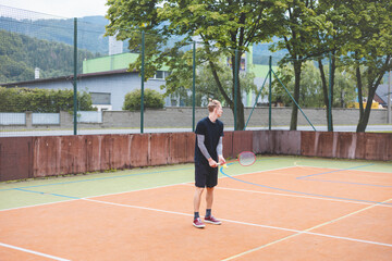 Fototapeta premium Young man prepares to play badminton on an outdoor court. Holding a racket and shuttlecock, he stands ready to serve in a casual setting surrounded by trees and urban elements