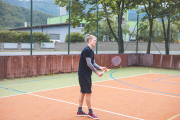 Fototapeta premium Badminton player stands ready to serve on an outdoor court, holding a racket and shuttlecock. Dressed in casual sportswear, he focuses on the game amid a scenic, natural setting