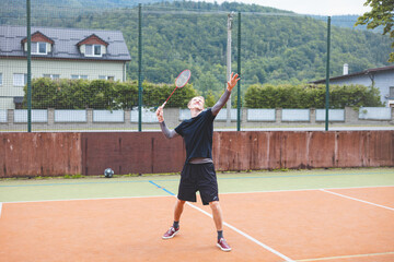 Badminton player stands ready to serve on an outdoor court, holding a racket and shuttlecock. Dressed in casual sportswear, he focuses on the game amid a scenic, natural setting