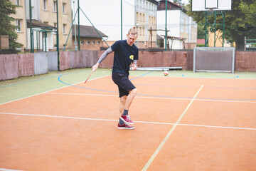Obraz premium Badminton player picks up a shuttlecock during a game on an outdoor court. The casual moment takes place in an urban setting, with surrounding buildings and a basketball hoop in the background