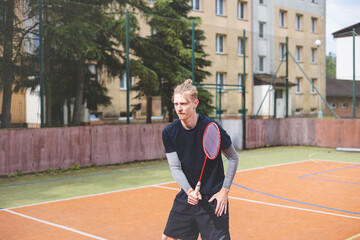 Badminton player focuses on the shuttlecock mid-air during a match on an outdoor court. With his racket raised, he prepares for the next shot, surrounded by an urban and scenic background