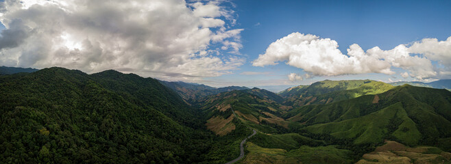 A rural road winds through a serene mountain vista, bordered by lush green forest under a blue sky...