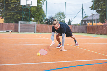 Obraz premium Badminton player stretches to retrieve a low shuttlecock during an intense game on an outdoor court. His athletic stance shows focus and determination, with an urban setting in the background