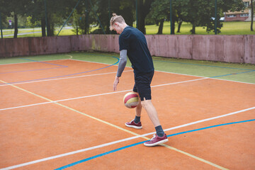 Young man dribbles a basketball on an outdoor court surrounded by nature. Dressed casually in sports attire, he showcases focus and skill during a casual practice session in a vibrant setting