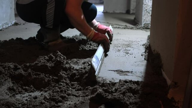 Builder worker making cement floor screed in rough finishing apartment. Repair master aligning fresh floor surface with spatula. Construction, overhaul, reconstruction work in unfinished building.
