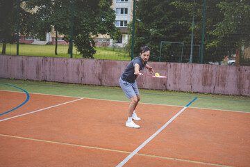 Badminton player focuses on the shuttlecock mid-air during a match on an outdoor court. With his racket raised, he prepares for the next shot, surrounded by an urban and scenic background
