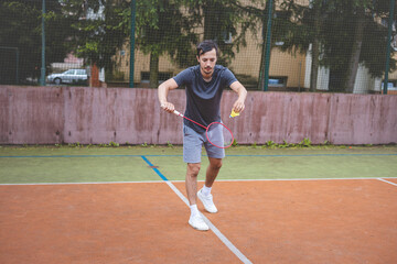 Badminton player stands ready to serve on an outdoor court, holding a racket and shuttlecock. Dressed in casual sportswear, he focuses on the game amid a scenic, natural setting