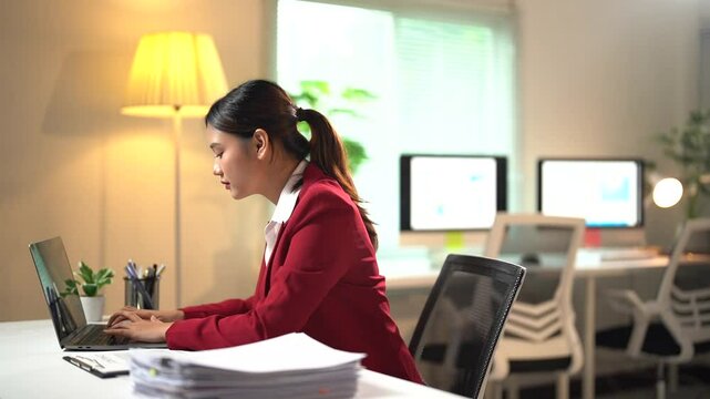 Businesswoman sits at her desk, working intently on her laptop. As time passes, fatigue sets in, causing her to slouch and hunch over while continuing to type