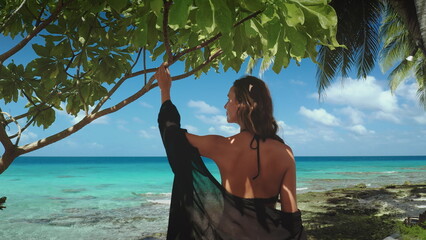 Young woman is admiring the turquoise blue lagoon of rangiroa island, french polynesia, sheltered from the sun by a tree