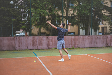 Obraz premium Badminton player strikes the shuttlecock during a game on an outdoor court, showcasing precise technique and athletic movement. The player is focused, surrounded by trees and an urban backdrop