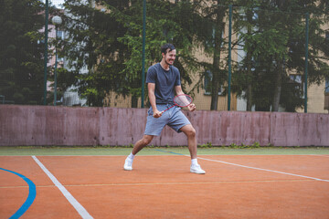 Obraz premium Badminton player celebrates a moment on an outdoor court, holding his racket in an energetic stance. His expression shows excitement and triumph, set against a backdrop of trees and buildings